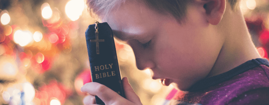 A young boy is holding a closed Holy Bible with a cross on its cover against his forehead. His eyes are closed, and he appears to be in deep prayer or reflection. The background features softly blurred, warm lights, adding a serene atmosphere to the scene—a moment of faith perhaps inspired by Vacation Bible School.
