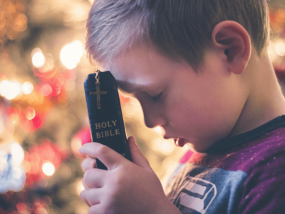 A young boy is holding a closed Holy Bible with a cross on its cover against his forehead. His eyes are closed, and he appears to be in deep prayer or reflection. The background features softly blurred, warm lights, adding a serene atmosphere to the scene—a moment of faith perhaps inspired by Vacation Bible School.