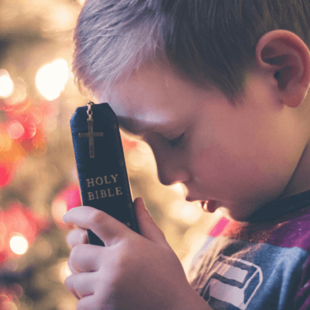 A young boy is holding a closed Holy Bible with a cross on its cover against his forehead. His eyes are closed, and he appears to be in deep prayer or reflection. The background features softly blurred, warm lights, adding a serene atmosphere to the scene—a moment of faith perhaps inspired by Vacation Bible School.