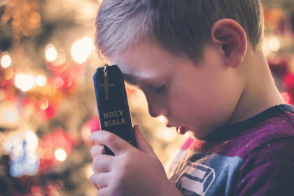 A young boy is holding a closed Holy Bible with a cross on its cover against his forehead. His eyes are closed, and he appears to be in deep prayer or reflection. The background features softly blurred, warm lights, adding a serene atmosphere to the scene—a moment of faith perhaps inspired by Vacation Bible School.